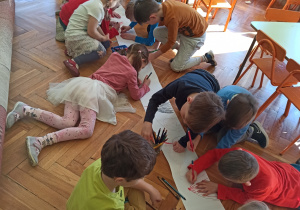 dzieci na papierze z długiej rolki wspólnie rysują " Świat dinozaurów". Praca odbywa się na podłodze